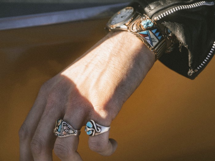 A man's arm hanging outside a car window, wearing a colorful watch and a couple of rings 